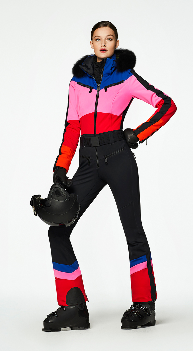 Model wearing a pink, red, blue and black color-block ski suit, holding a black ski helmet