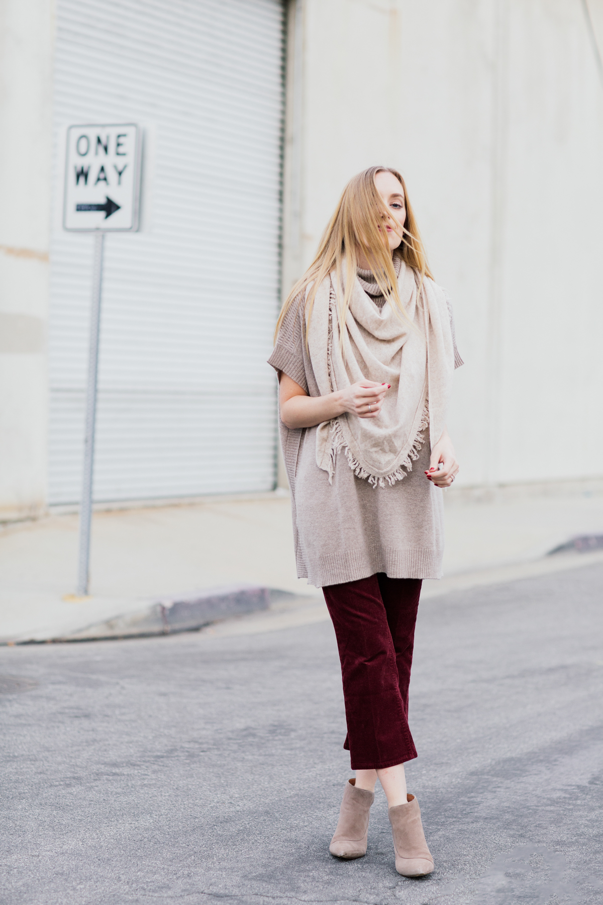 Woman in a fringed beige triangle scarf, taupe poncho knit, burgundy corduroy trousers and beige suede booties