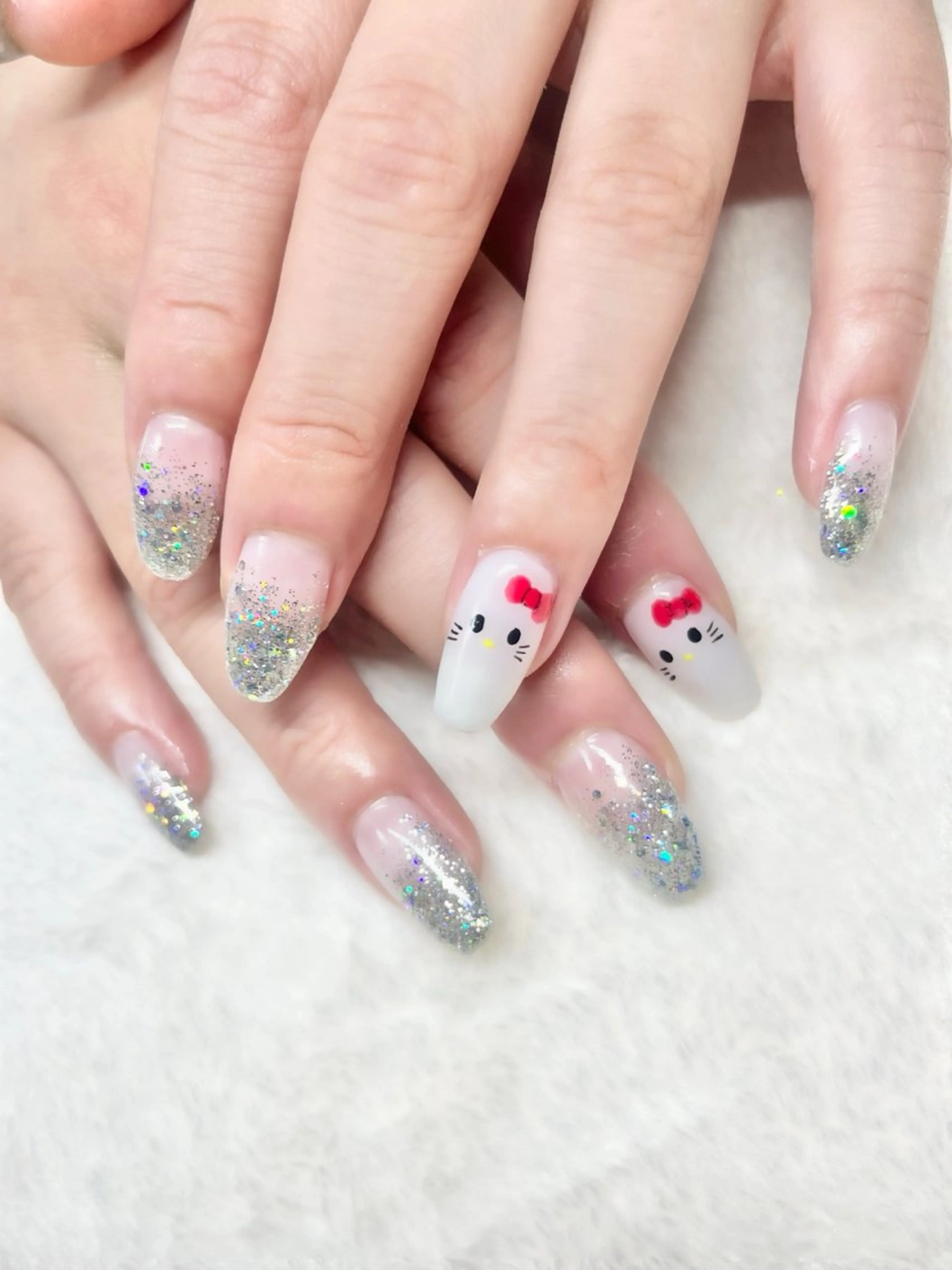 Medium almond nails in sheer nude with silver holographic glitter ombre tips and simple white Hello Kitty faces with red bows