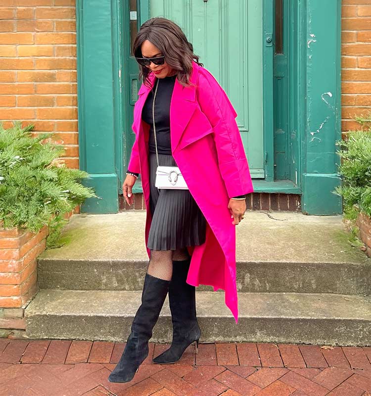 Woman in a fuchsia long coat with a black top, pleated skirt and knee-high boots