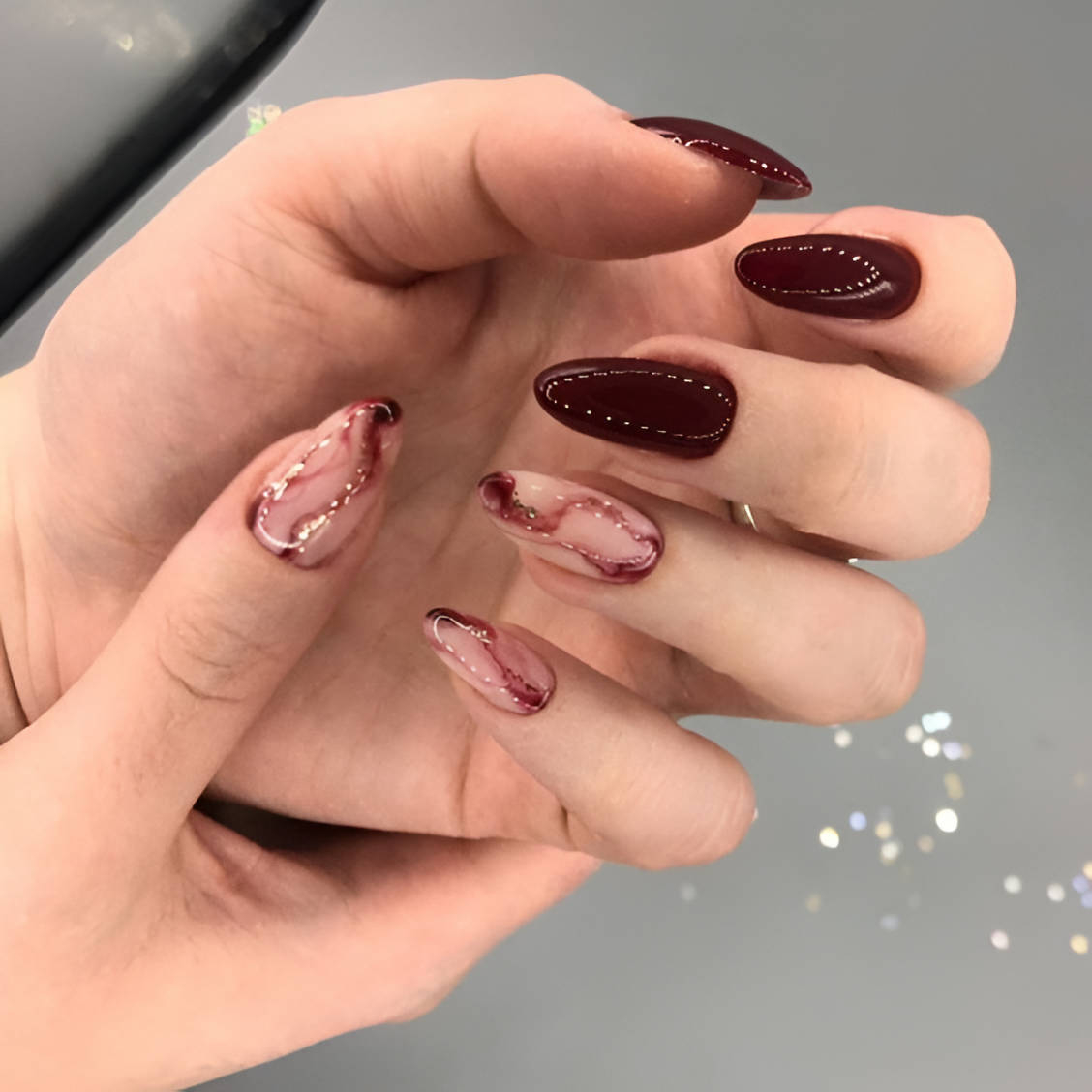 Almond nails in deep maroon with two blush-pink marble accent nails veined with gold glitter; real photography.