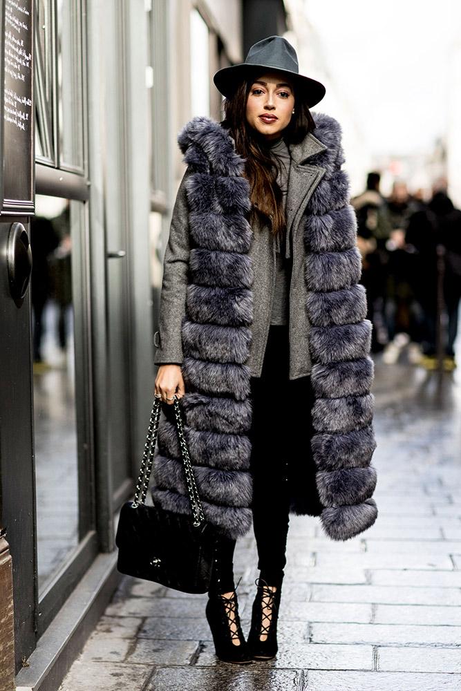 A woman wearing a long grey faux-fur vest over a blazer with black skinny pants and a fedora