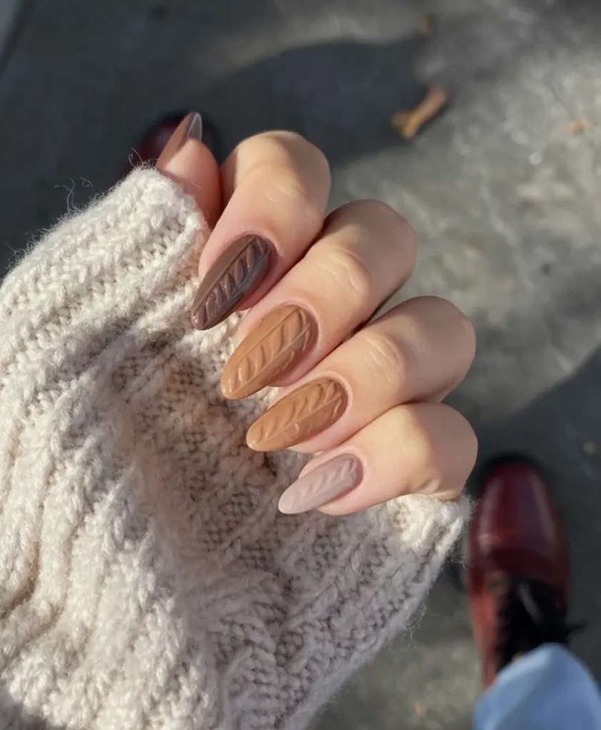 Almond nails in chocolate brown and mocha shades with embossed cable-knit sweater texture; the hand is tucked into a cream cable-knit sweater cuff. Real candid photo, no watermark. (A woman-trend.com alternative was rejected as AI-suspect.)