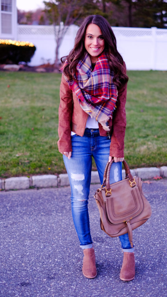 Woman in a rust suede moto jacket with a red plaid blanket scarf, ripped jeans, and tan ankle boots