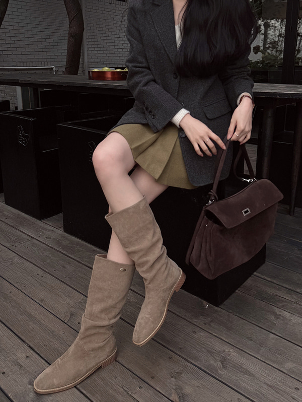 Woman seated wearing taupe slouchy flat boots with a khaki mini skirt, dark blazer and brown bag
