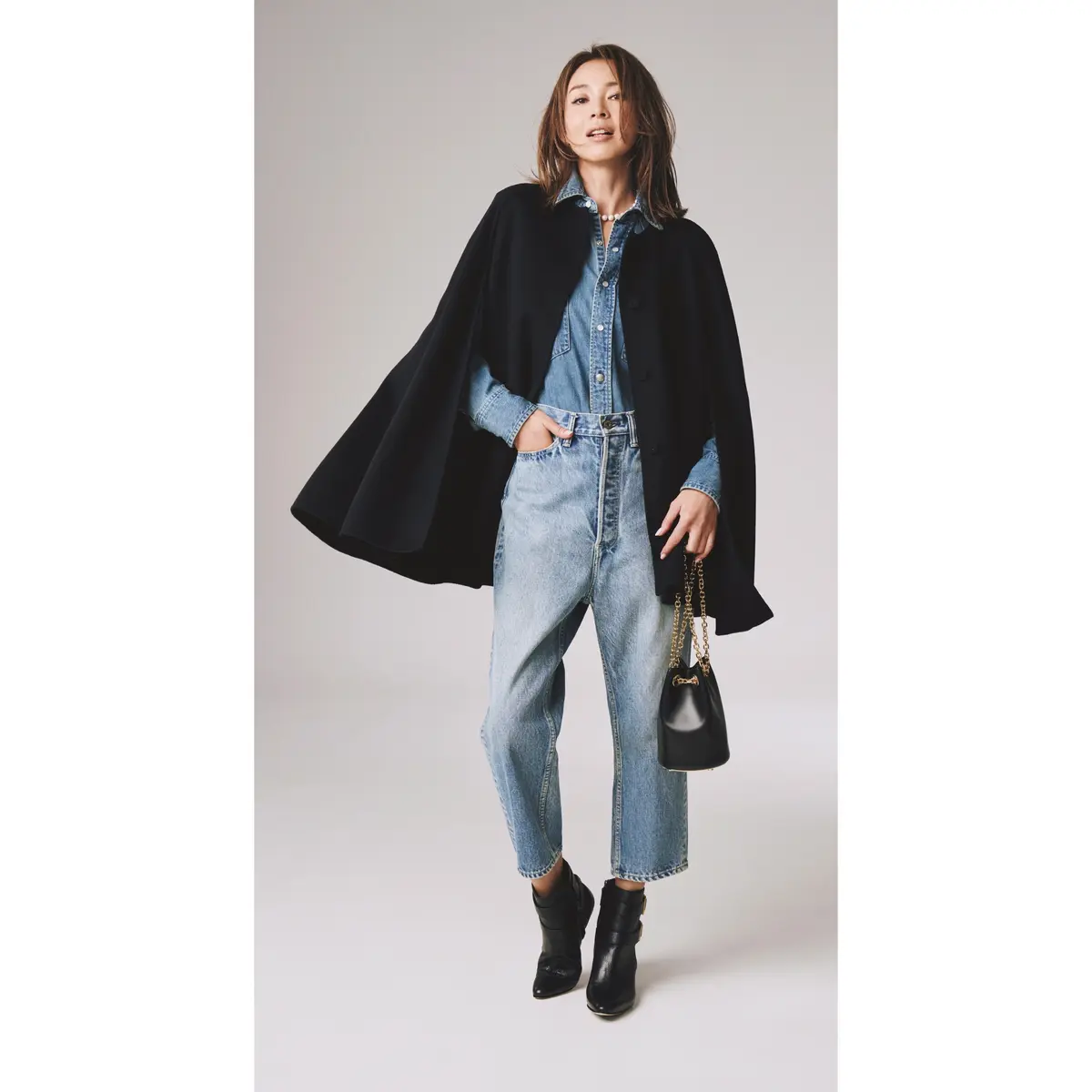 Woman wearing a black cape coat over a denim shirt and light-wash jeans with black ankle boots