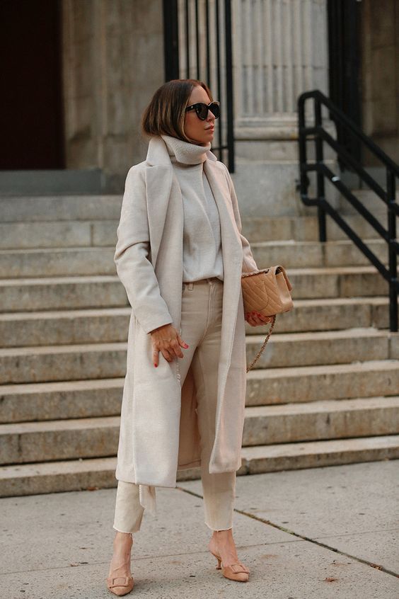 Woman in a long ivory wool coat, cream turtleneck and cream trousers with nude heels