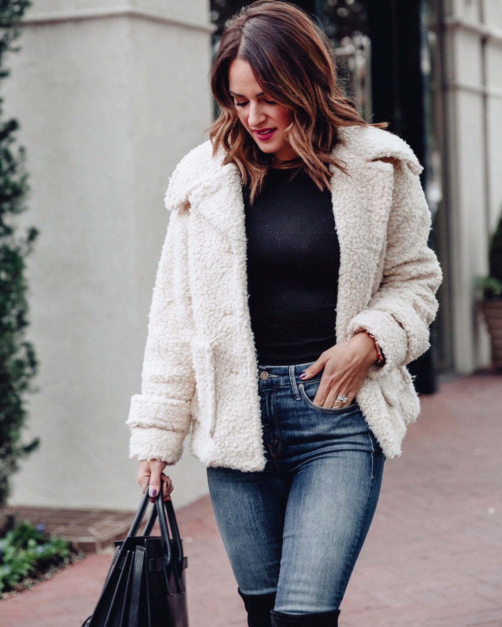 Woman in a cream sherpa jacket over a black knit top with skinny jeans and a black tote
