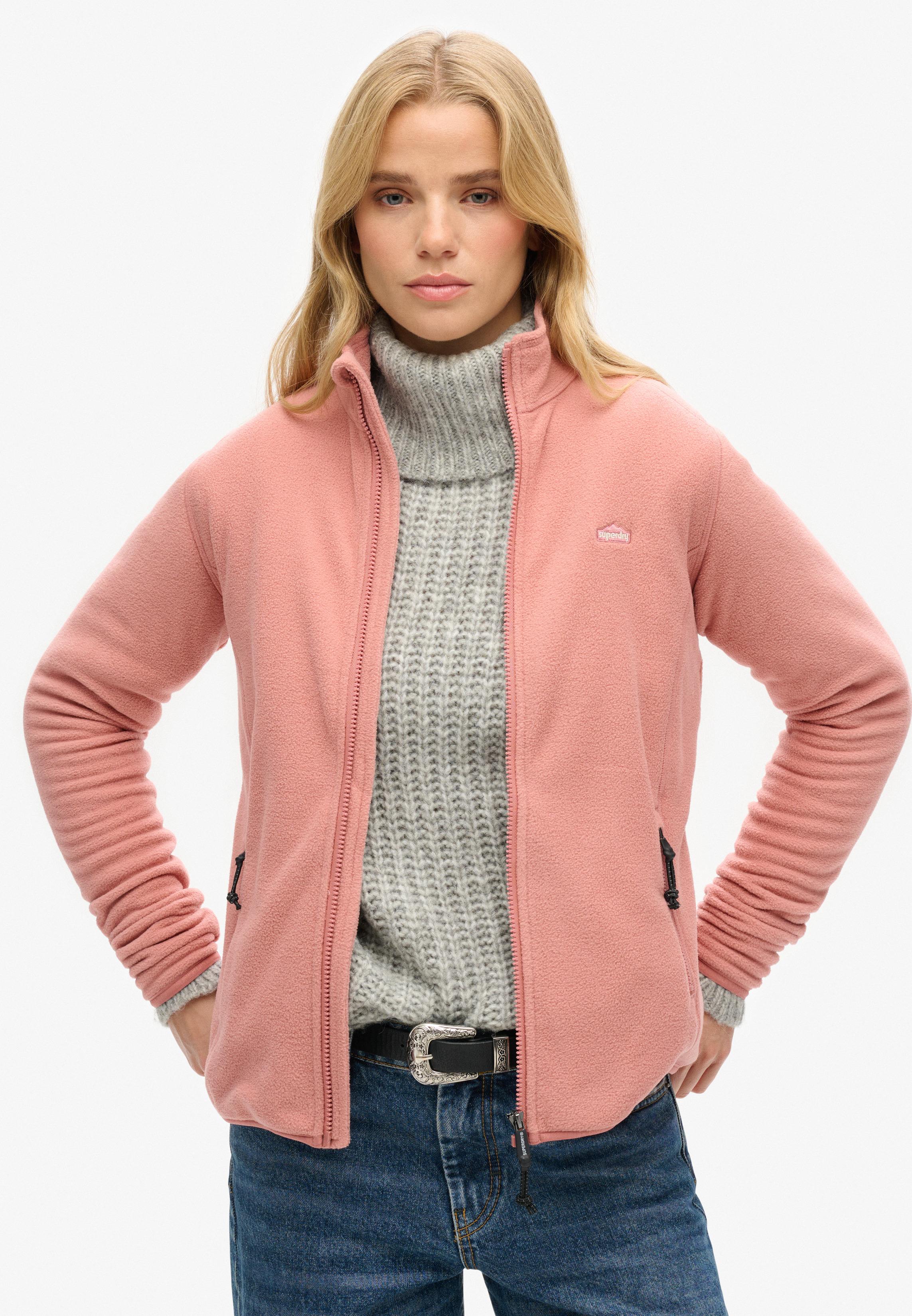 Woman in a pink fleece jacket over a grey chunky turtleneck with jeans and a statement belt