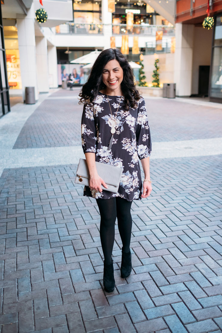 Woman in a dark floral mini dress with black tights, black ankle boots and a grey clutch