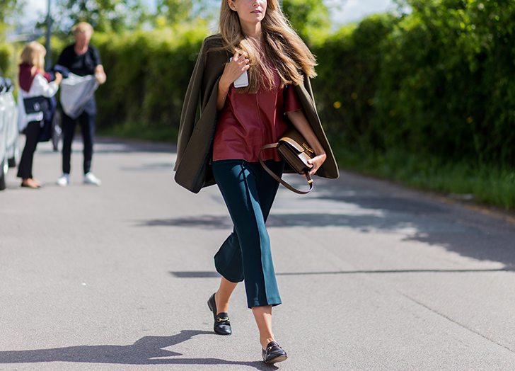 Woman wearing a rust blouse and teal cropped trousers with black loafers, an olive coat draped over her shoulders and a brown bag