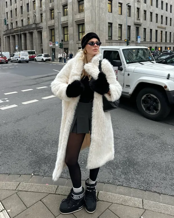 Woman in a long white teddy coat with a pleated mini skirt and snow boots in the snow