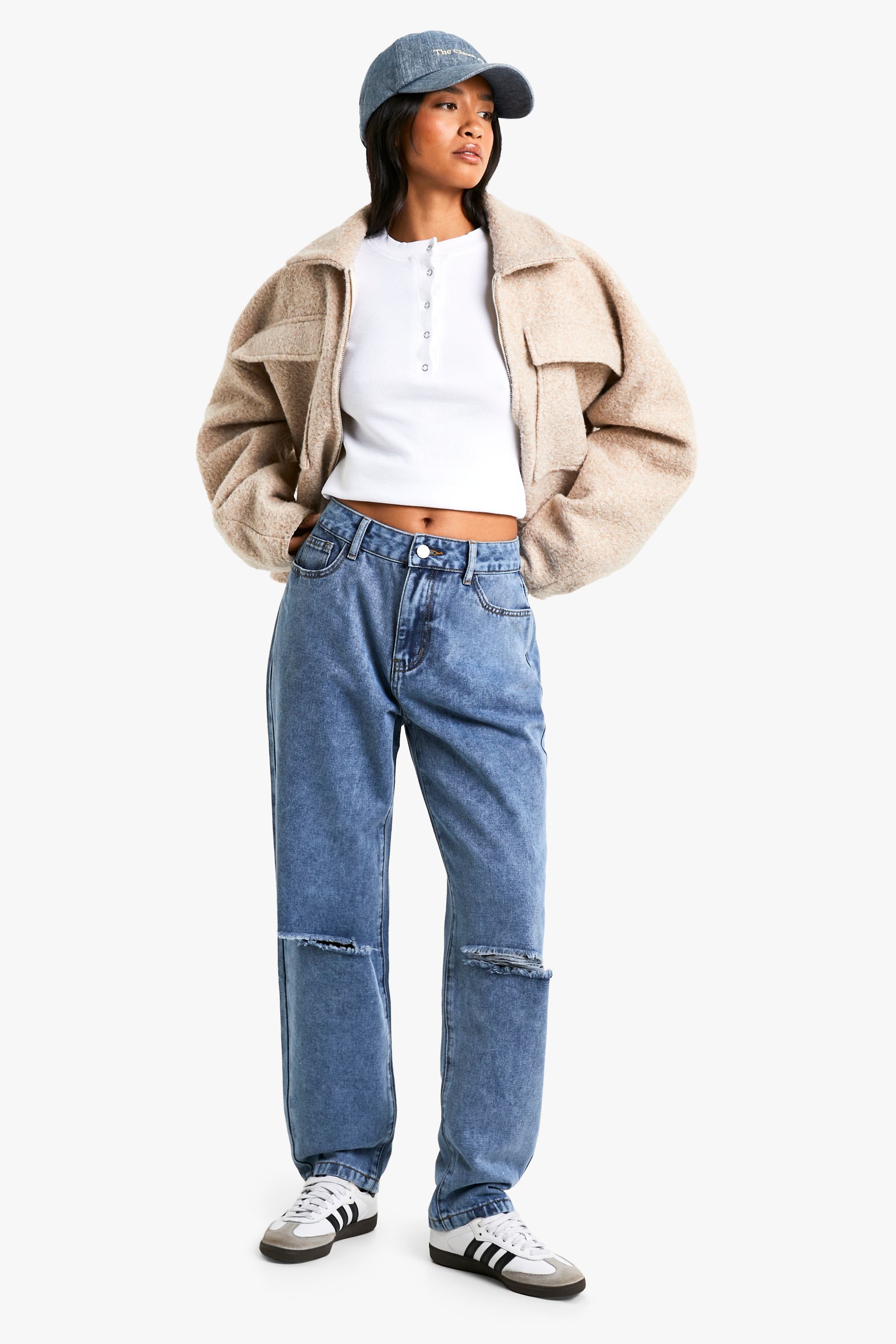 Woman in a beige sherpa trucker jacket with a white henley, ripped jeans, sneakers and a denim cap