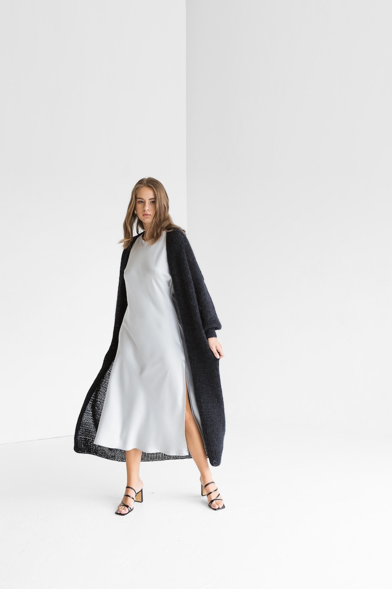Woman in a long dark mohair cardigan worn open over a white satin slip dress with strappy heeled sandals
