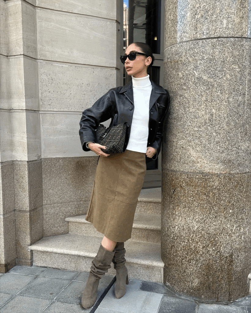 Woman in taupe slouchy heeled boots with a brown suede midi skirt, white turtleneck and black leather jacket