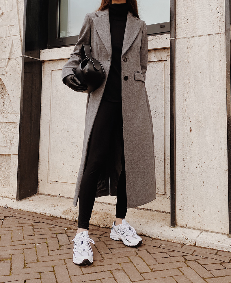 Woman in a long grey coat, black turtleneck, black leggings and white sneakers