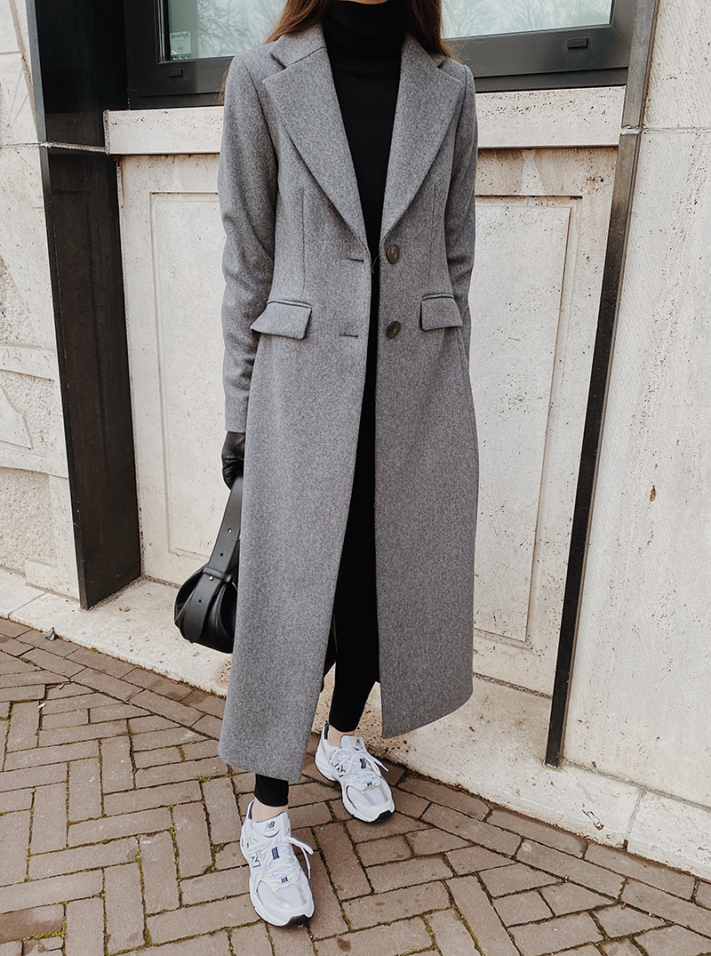 A light grey long coat over a black turtleneck and leggings with white sneakers and a dark bag.