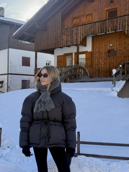Woman in a cinched black puffer jacket with a grey scarf and sunglasses in front of a wooden chalet