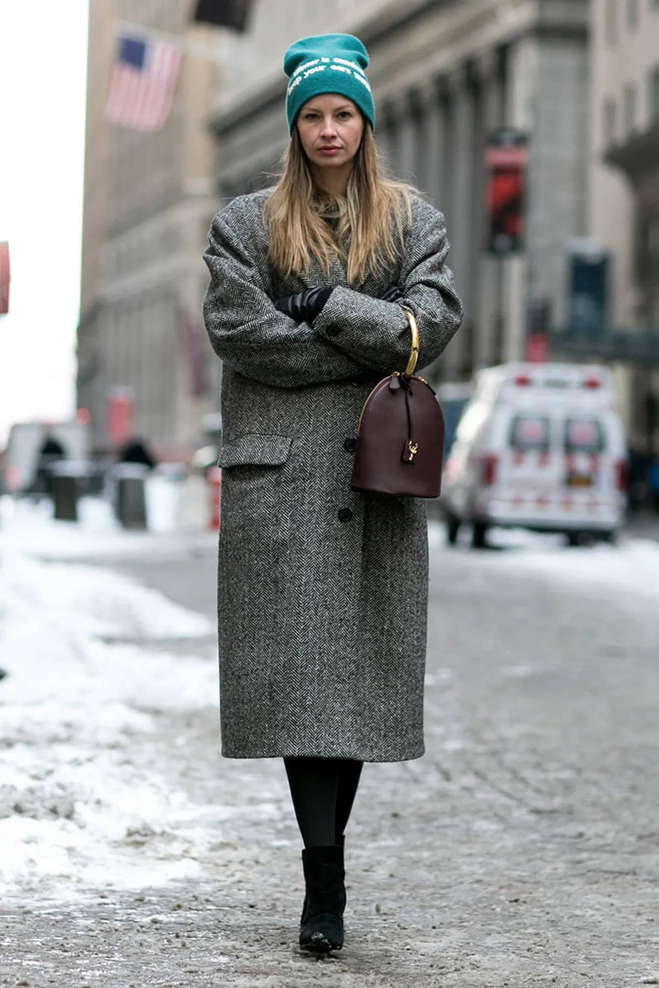 Woman in a teal logo beanie and long herringbone coat on a snowy street