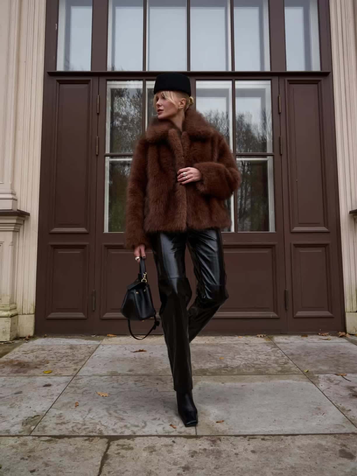 Woman in a brown faux-fur jacket, black cap, black shiny trousers and ankle boots holding a black bag