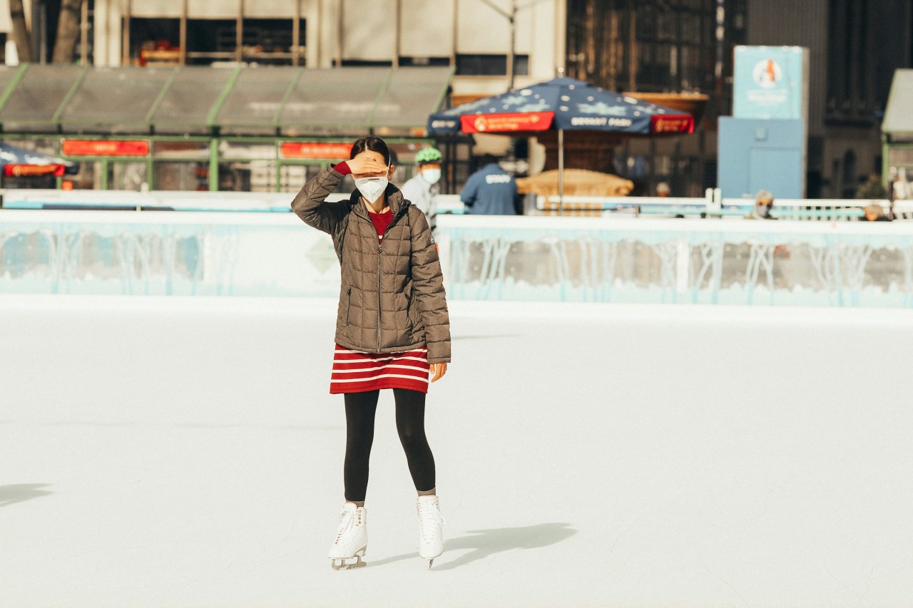 Woman on an outdoor ice rink in a grey quilted puffer, red striped skirt, black leggings and white skates