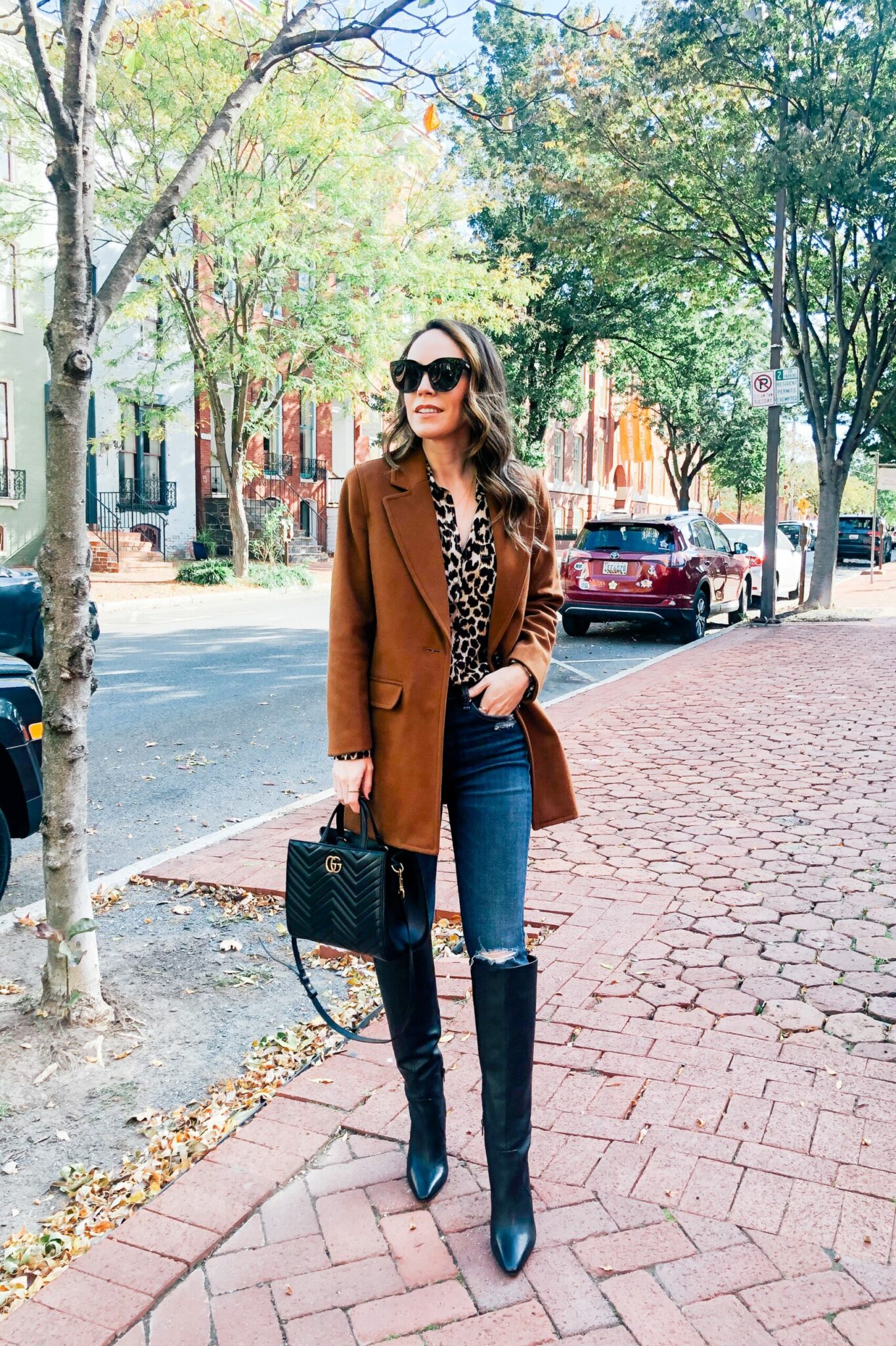 Woman in a camel coat over a leopard-print blouse and dark jeans with knee-high black boots