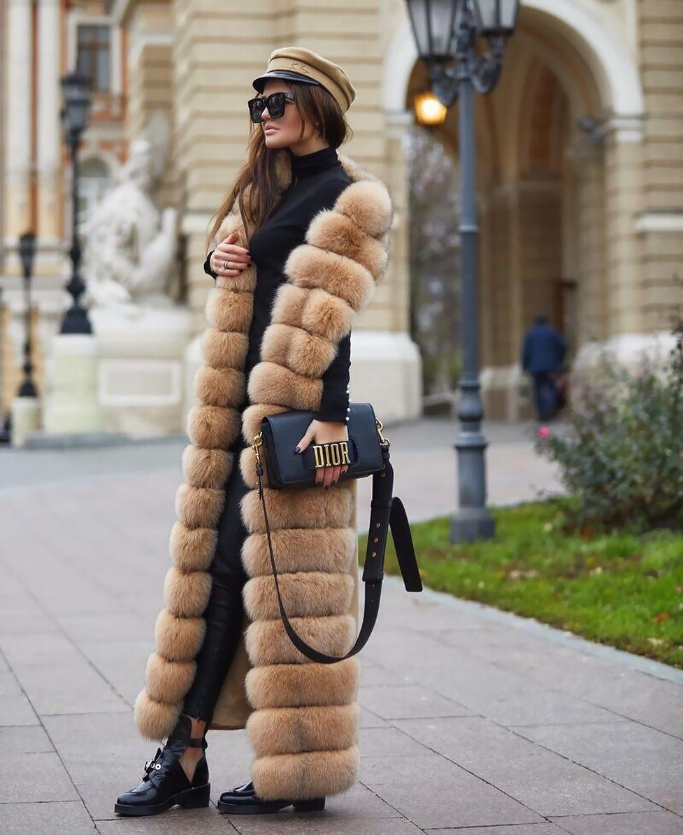 A long camel faux fur vest with horizontal fur bands, worn open over a black turtleneck and black trousers, finished with a baker boy cap and a structured black bag.