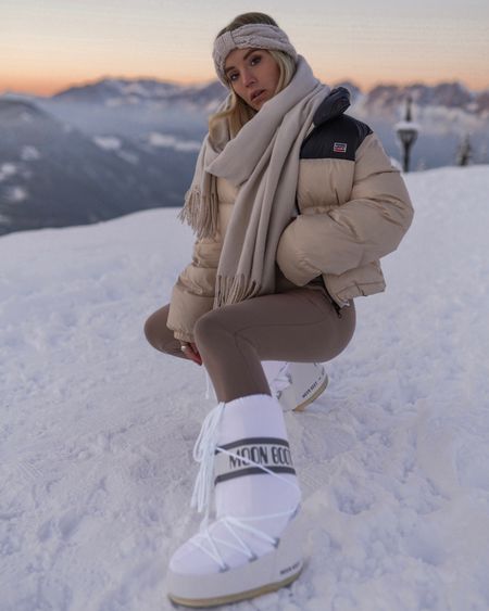 Woman in white Moon Boots with logo band, beige puffer, beige scarf and taupe leggings crouching in snow at sunset