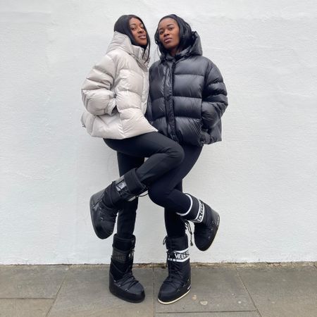Two women in black moon boots, cropped puffers and black leggings posing together against a white wall