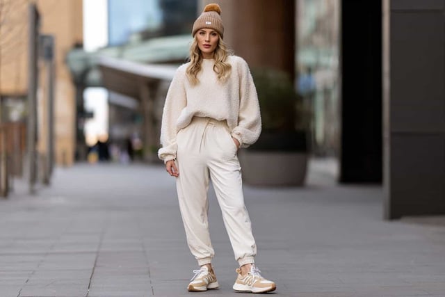 Woman in a cream textured sweater and white joggers with a tan beanie and sneakers outdoors
