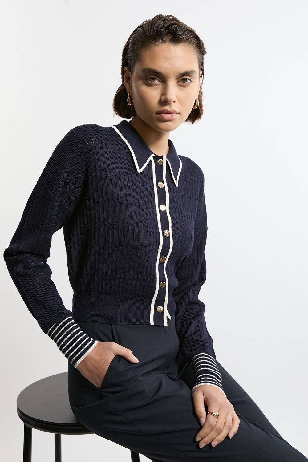 Woman wearing a navy collared cardigan with white trim and gold buttons with navy trousers