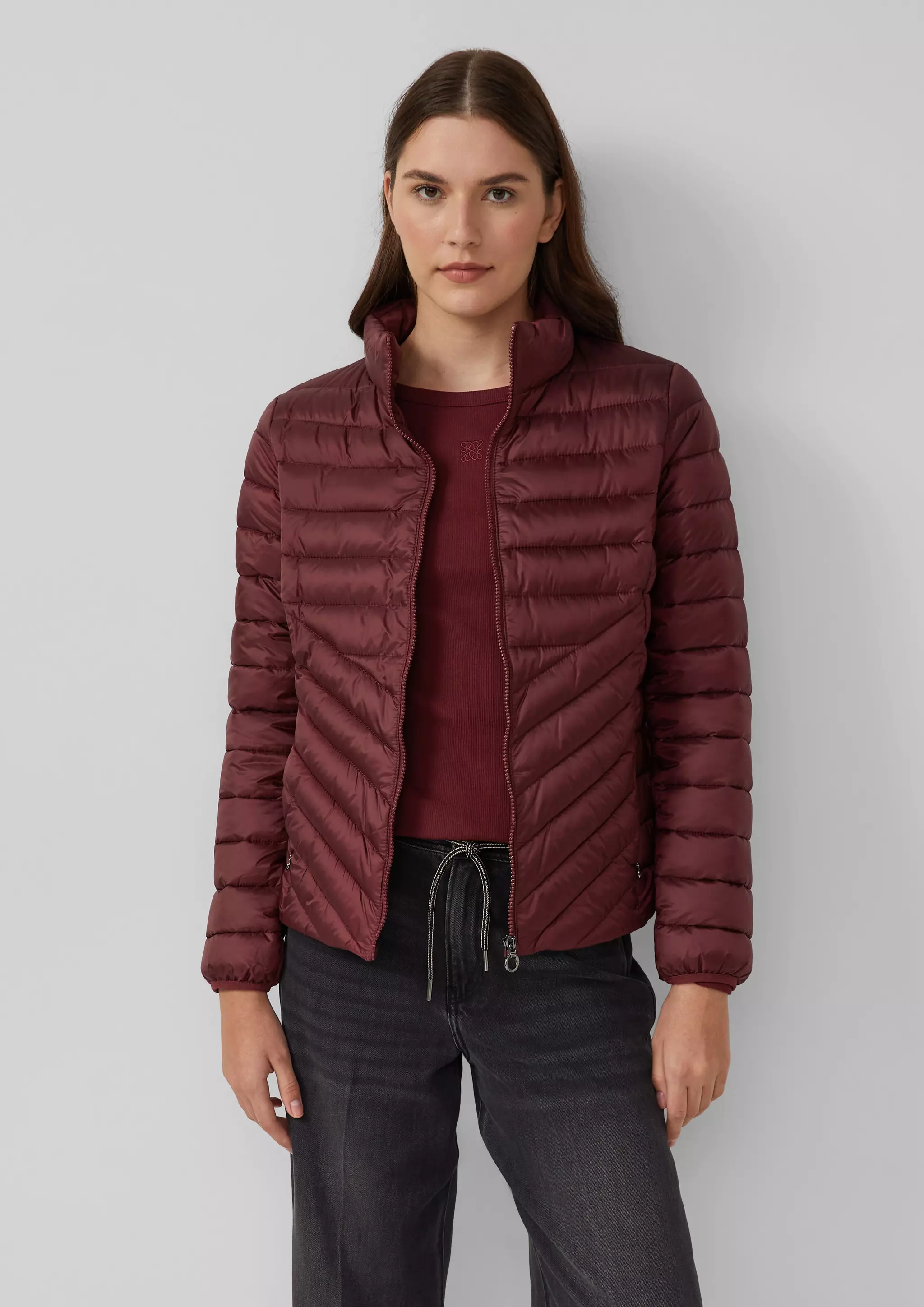 A lightweight burgundy quilted puffer jacket with chevron stitching, worn open over a matching burgundy top and dark grey jeans with a drawstring waist.