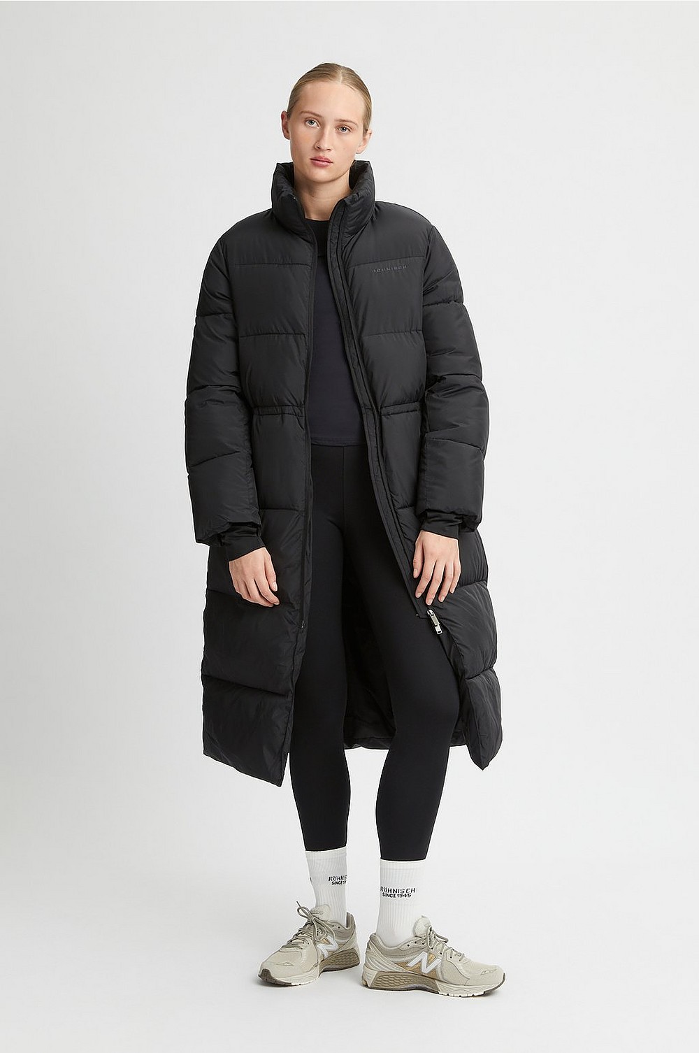Woman in a long black puffer coat, black leggings, white socks and beige sneakers