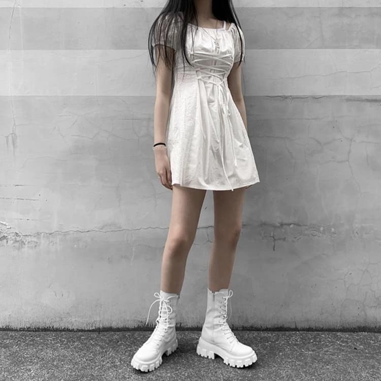 Woman in a white ruched mini dress and white chunky platform combat boots against a concrete wall