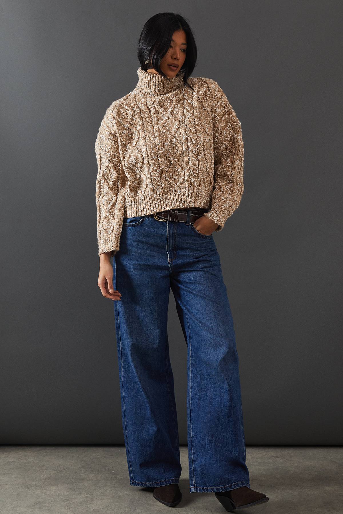 Woman in an oatmeal cable-knit turtleneck with medium-wash wide-leg jeans, a brown belt and western boots