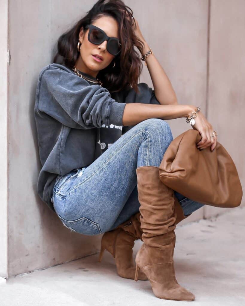 Woman crouching in tan slouchy heeled boots, distressed jeans and a grey sweatshirt