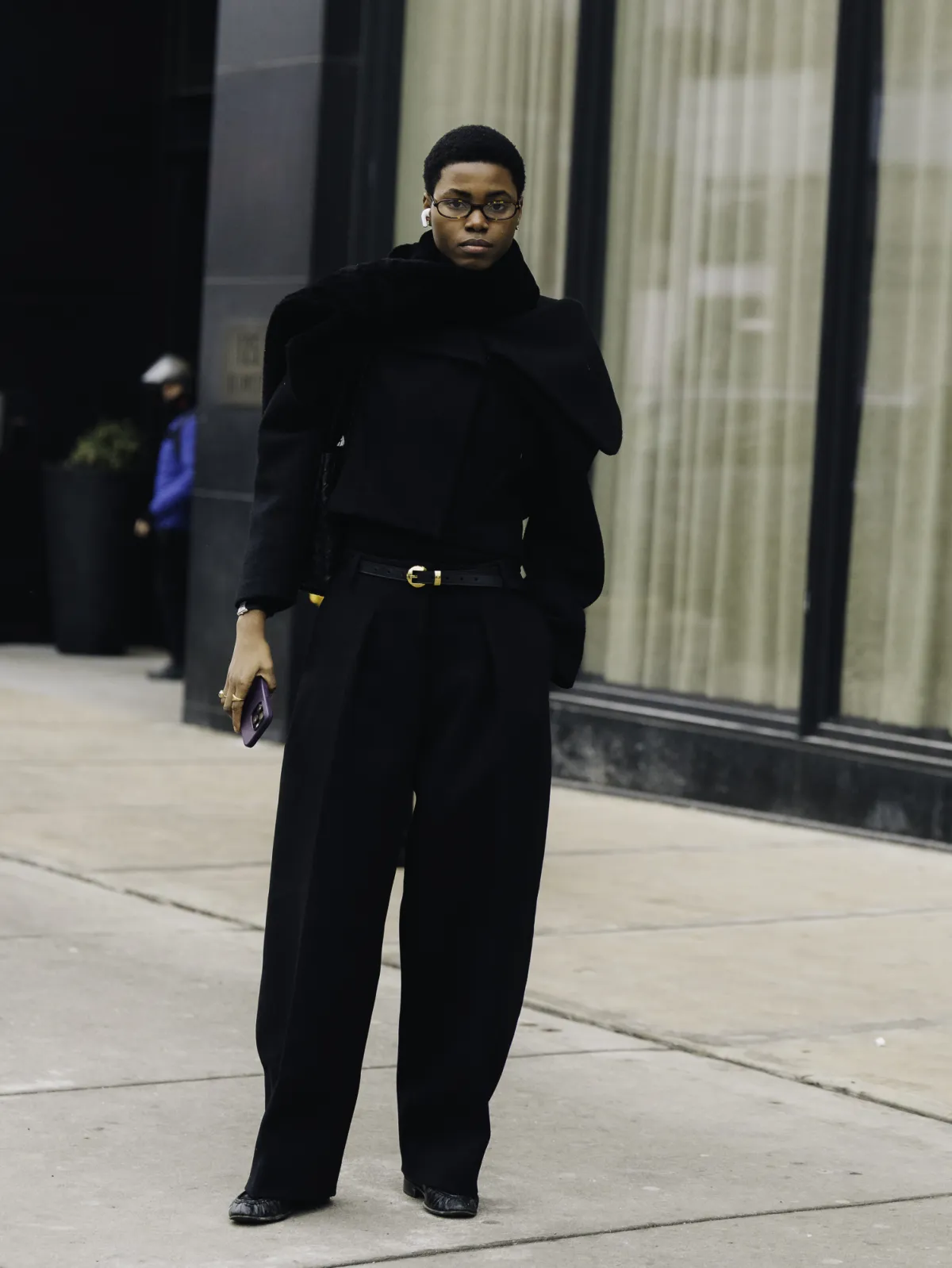 Woman in an all-black outfit with a draped black coat, wide-leg trousers and a gold-buckle belt at New York Fashion Week