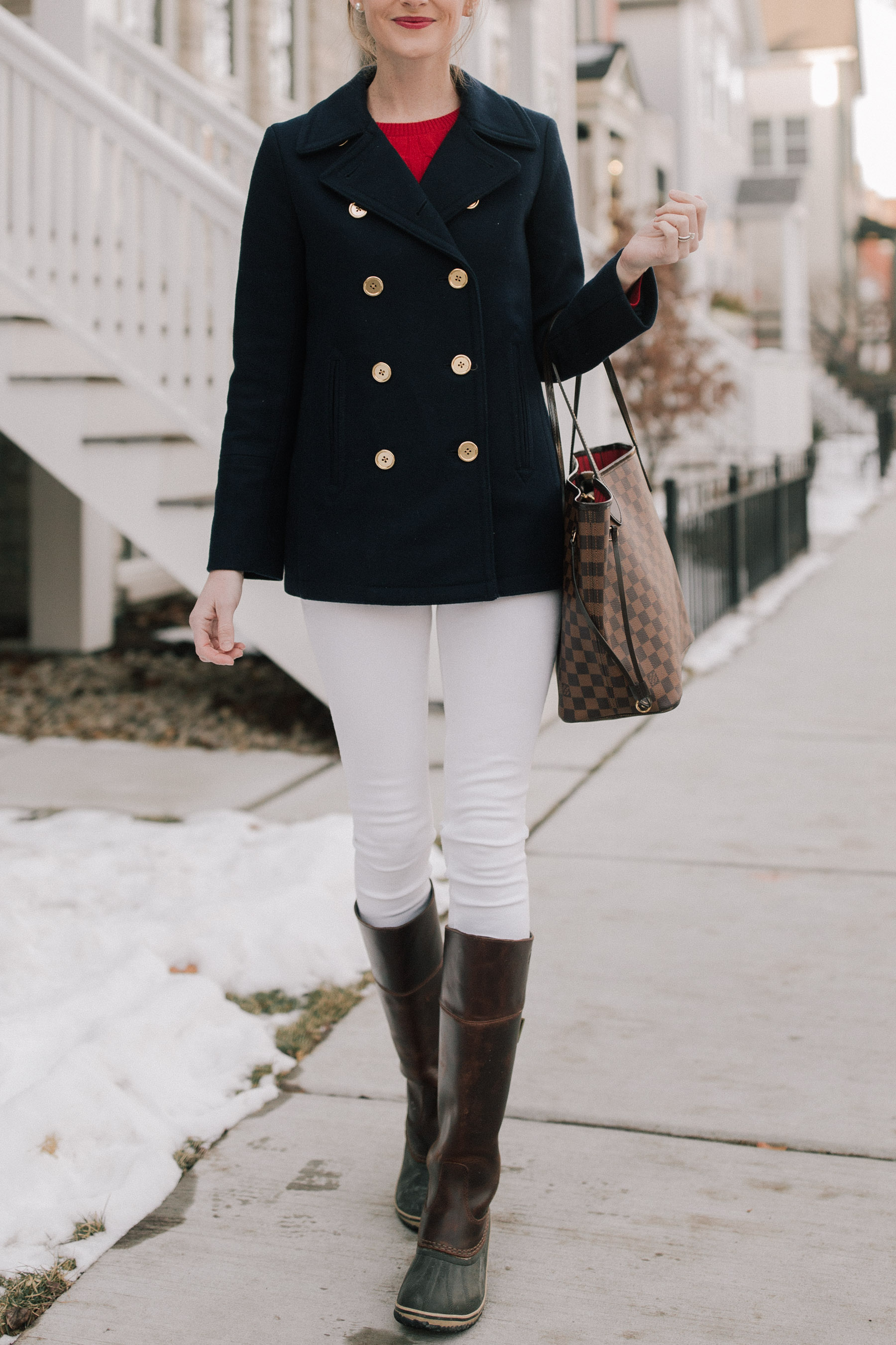 Woman in a navy double-breasted peacoat with white skinny jeans tucked into tall brown boots, carrying a checker tote