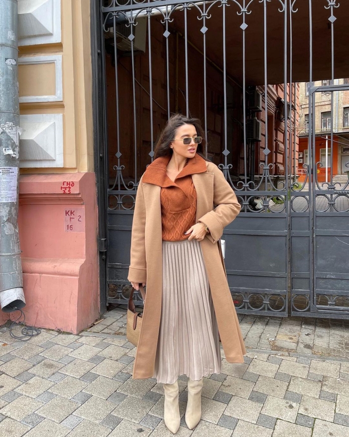 Woman in a camel coat over a beige pleated midi skirt and a cable-knit sweater with beige ankle boots