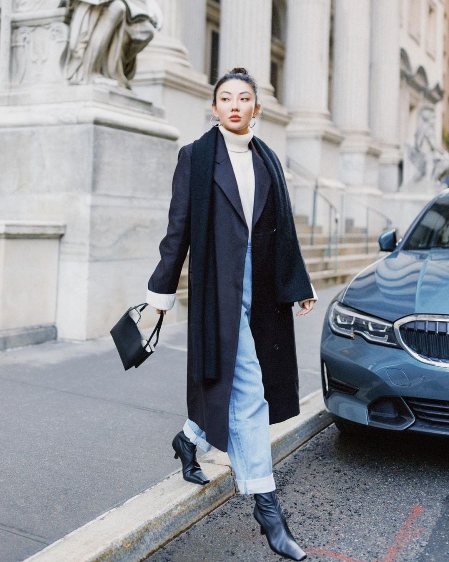 Woman wearing a black wrap coat over a white turtleneck and light-wash jeans with black ankle boots