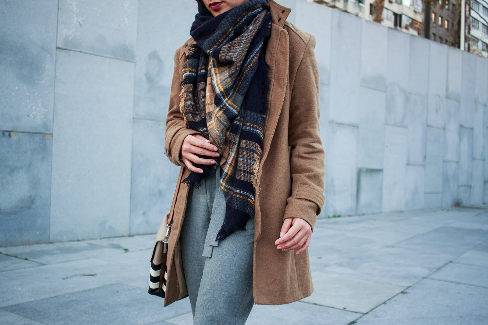 Woman in a tan coat with a large navy and mustard tartan blanket scarf and grey trousers