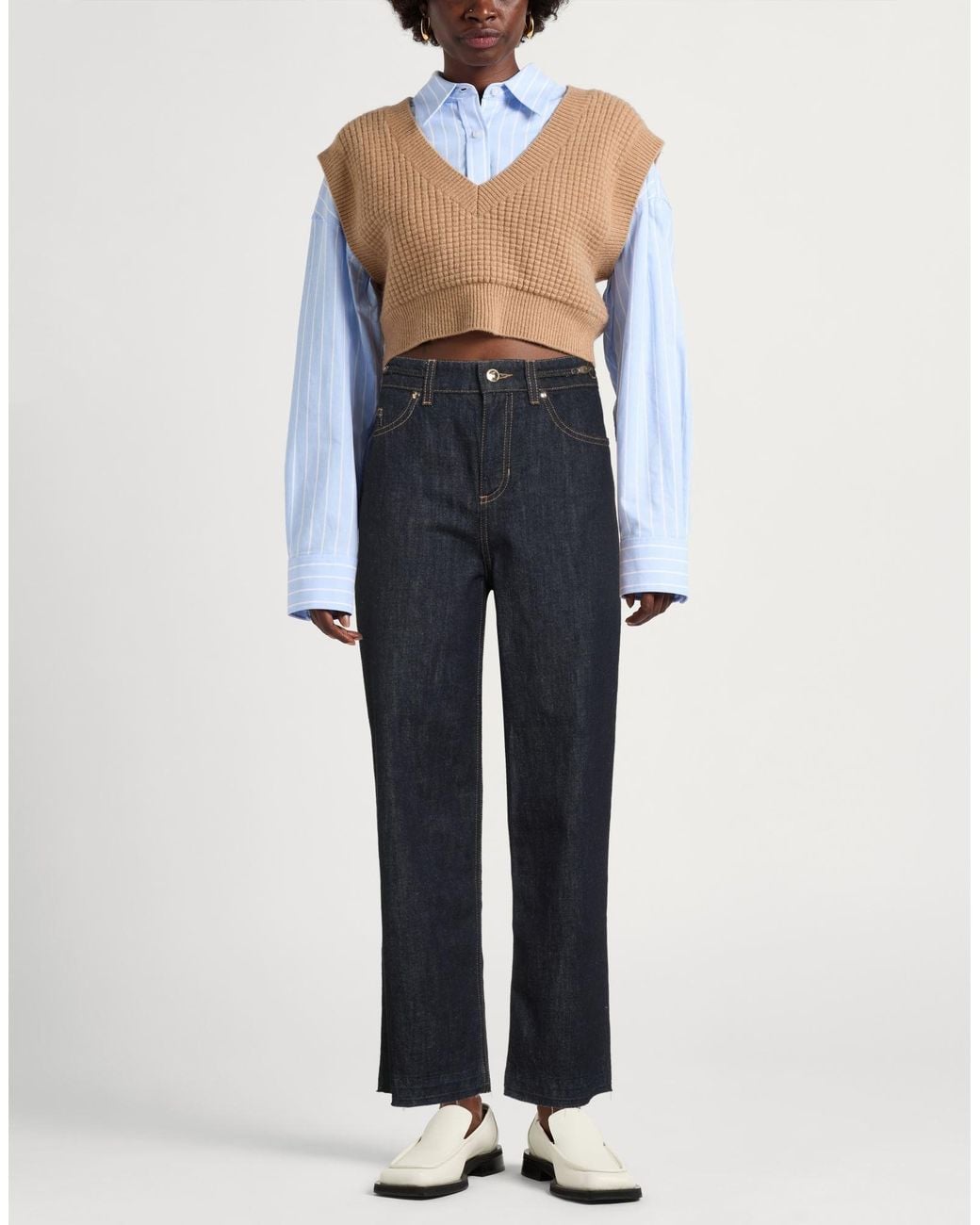 Woman in a tan cropped knit vest over a striped blue shirt with dark wide-leg jeans and white loafers