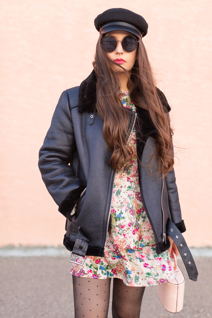 Woman in a black shearling jacket with a floral mini dress, polka-dot tights and a baker-boy cap