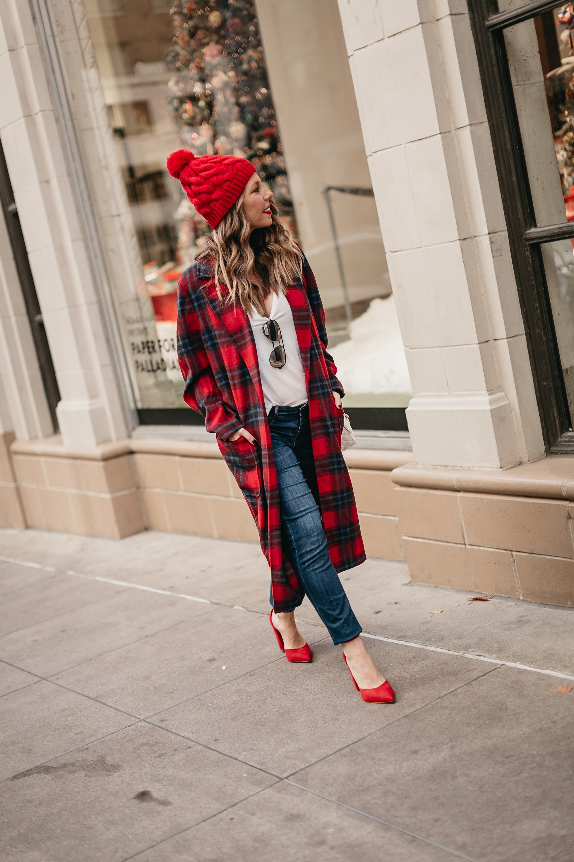 Woman in a red and black plaid long coat with a red pom beanie, white top, jeans and red heels