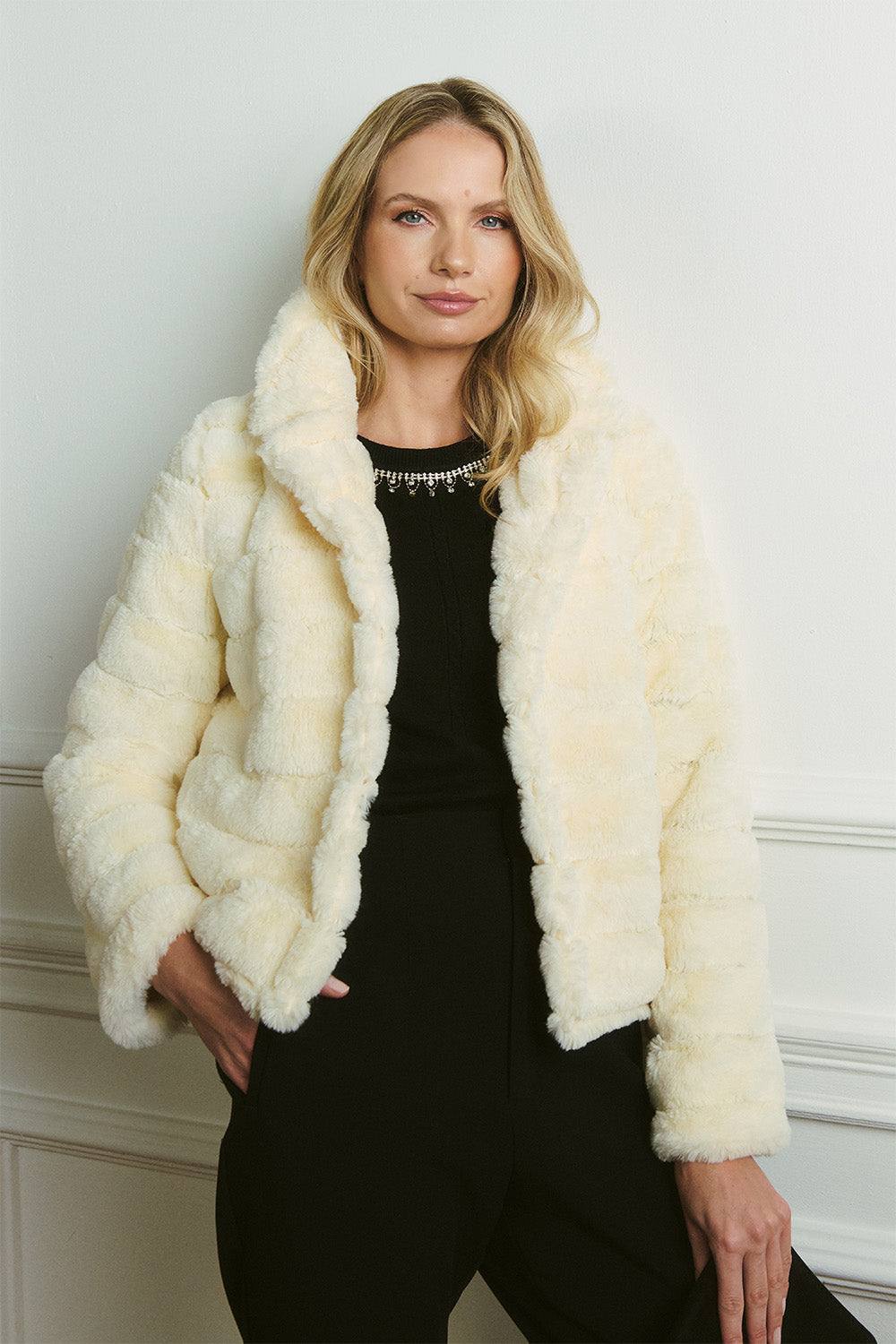 A cream quilted faux fur cropped jacket worn open over a black top with an embellished neckline and black trousers.