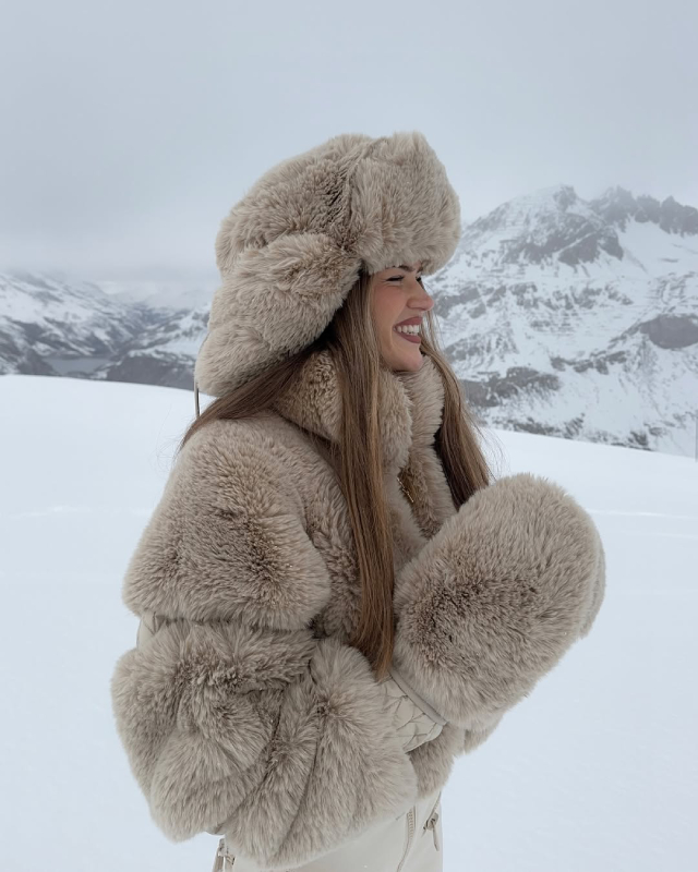 Woman in a beige faux fur trapper hat and beige fur jacket with snowy mountains behind