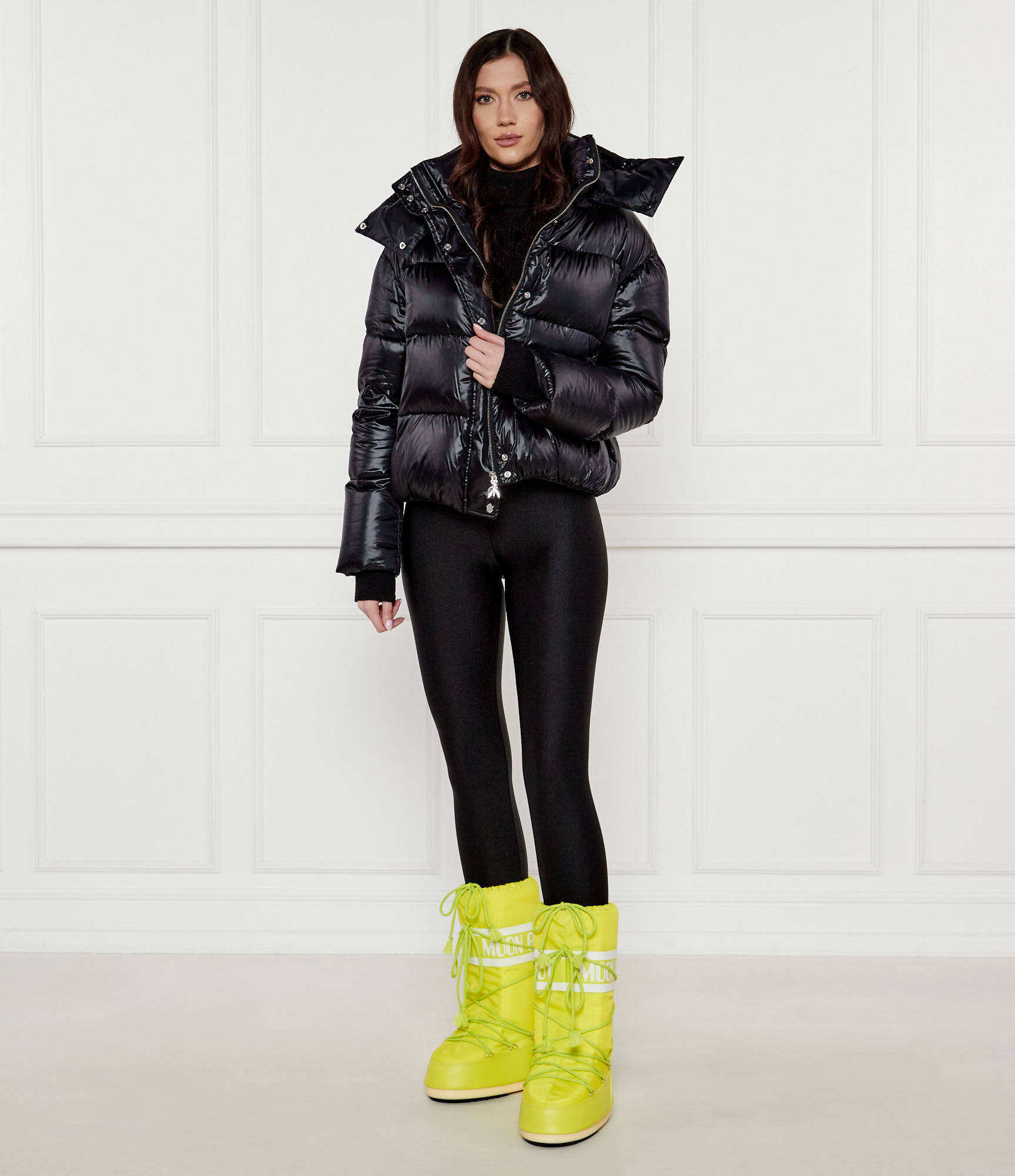 Woman in neon lime green Moon Boots, black glossy puffer jacket and black leggings in a studio