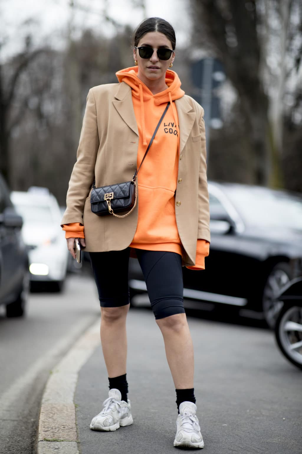 Woman in orange hoodie, camel blazer, black bike shorts and chunky sneakers