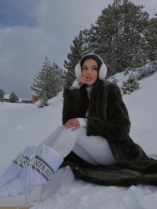 Woman in an olive faux-fur coat, white leggings and white moon boots sitting in the snow
