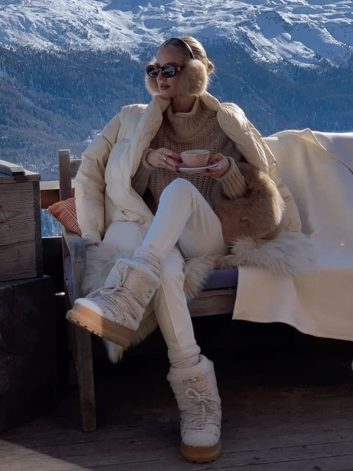 Woman in cream furry moon boots, cream puffer with fur collar, brown knit and white pants on an alpine terrace